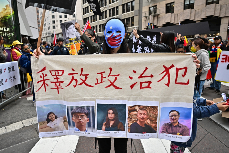 2023年11月17日，在習近平下榻的舊金山飯店附近，一名戴著繪有東突厥斯坦旗幟面具的抗議者，手持要求釋放政治犯的布條──包括在微信分享白紙革命影片的維吾爾學生卡米萊．瓦依提、發起北京四通橋抗議的彭立發，以及因香港民主派47人初選等案被押的黃之峰、何桂藍、戴耀廷。（攝影／AFP／Frederic J. Brown）