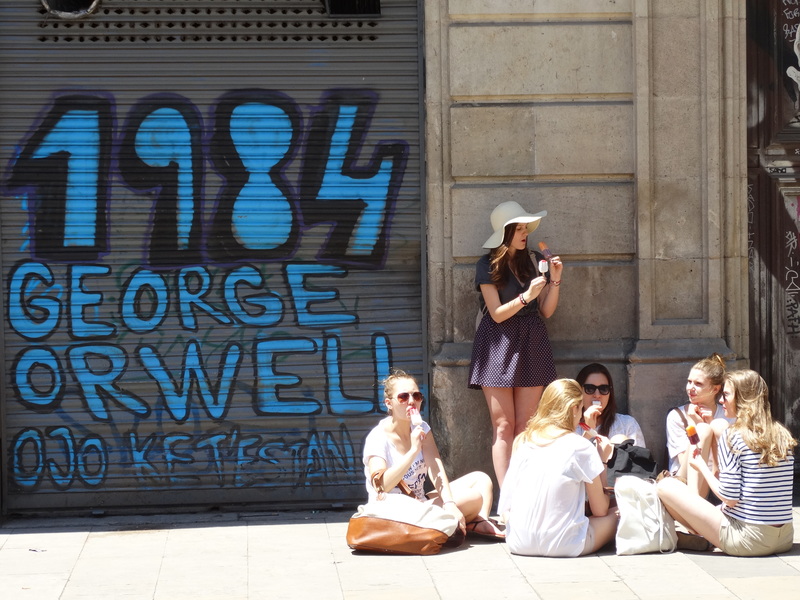 2014年6月1日，位於西班牙巴賽隆納的喬治・奧威爾廣場（Placa de George Orwell），觀光客在一處有「1984」塗鴉的鐵捲門邊休息。（攝影／Adam Jones／Wikimedia Commons - CC BY-SA 2.0）