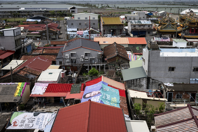 2025年7月31日，台南七股的頂山里，從高處往下望，許多曾經的紅瓦屋頂被印著政治人物臉孔與建商廣告的帆布覆蓋，因屋瓦嚴重缺料或停產，在短期內修復極爲困難，部分居民已將屋頂換成紅色或灰色鐵皮。這場風災不僅打擊民生，也永遠改變了溪北地區地景。（攝影／楊子磊）
